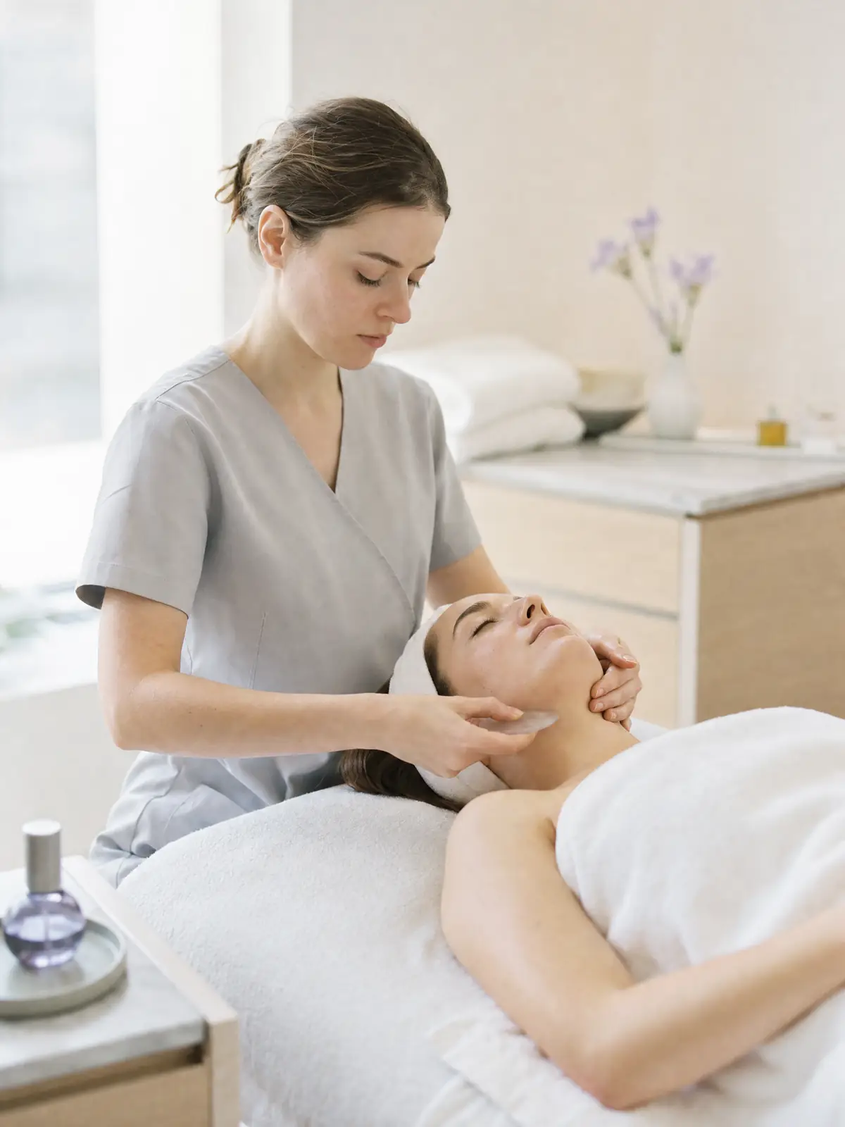 An esthetician working a treatment along a reclining client's jawline in a serene, minimal room