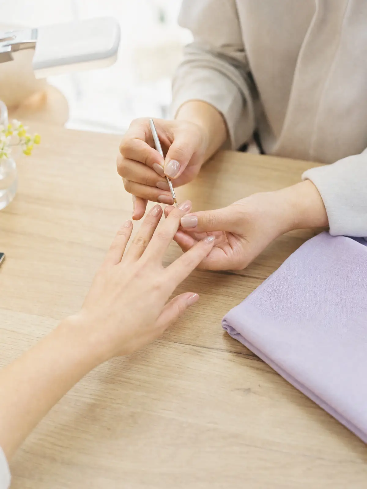 A nail technician's hands finishing a glossy neutral gel manicure at a pale wood table