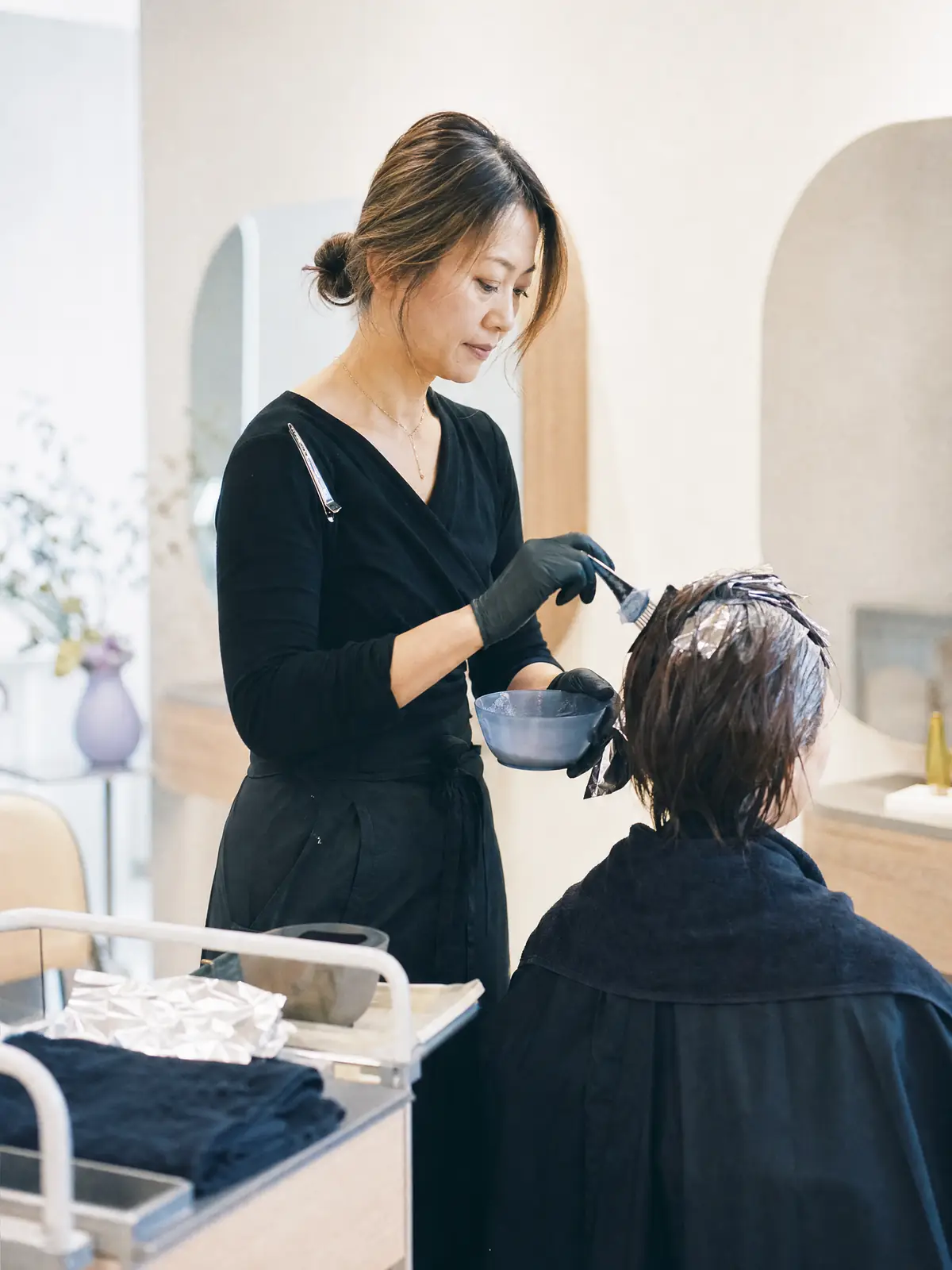A stylist sectioning and painting colour into a client's hair at a sunlit mirror station